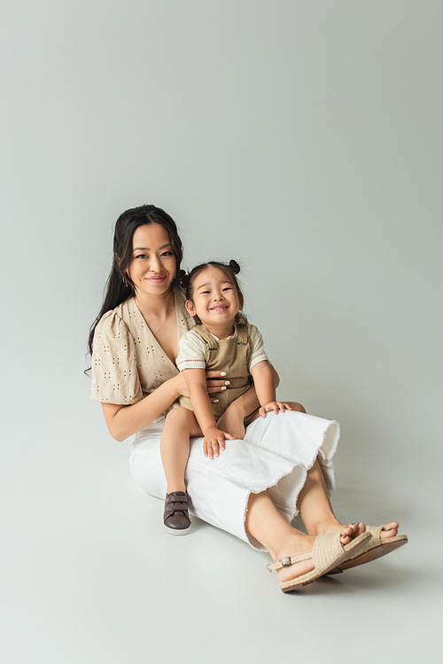 full length of cheerful asian mother sitting with toddler daughter on grey