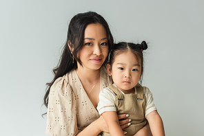 cheerful asian woman hugging toddler daughter isolated on gray