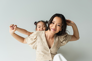 cheerful asian woman holding hands with toddler daughter isolated on gray