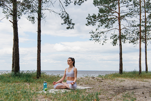 brunette young woman in sportswear sitting in lotus pose and meditating on yoga mat