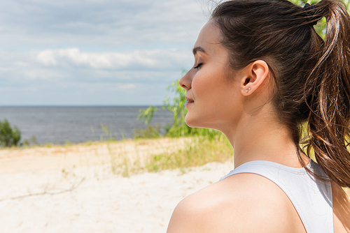 young and brunette woman with closed eyes meditating near ocean
