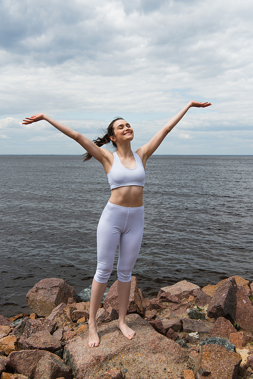 happy young woman with closed eyes and raised hands near sea