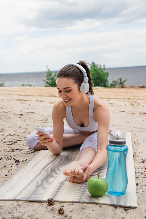 smiling woman in wireless headphones listening music while stretching on yoga mat and using smartphone