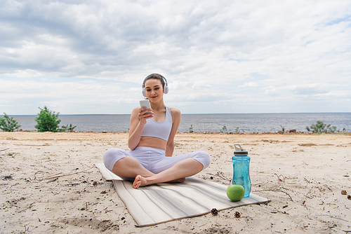 smiling woman in wireless headphones listening music while using smartphone and sitting with crossed legs on yoga mat