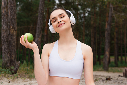 happy woman in headphones listening music while holding apple outside