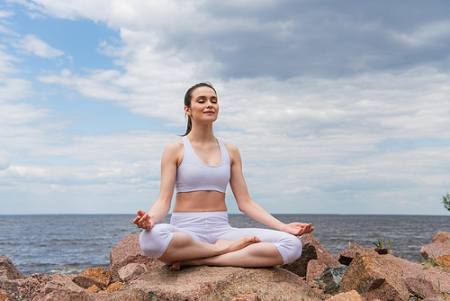 cheerful woman in headphones and sportswear sitting in lotus pose while meditating near sea