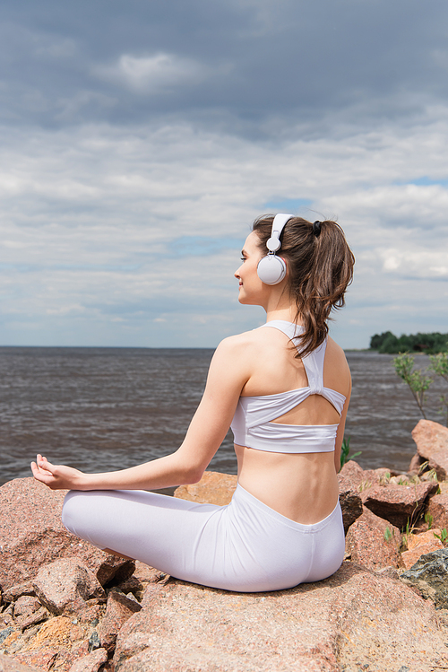 happy woman in headphones and sportswear sitting in lotus pose while meditating near sea