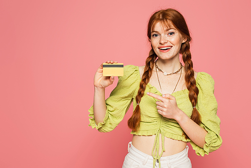 Smiling redhead woman pointing at credit card isolated on pink