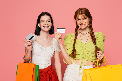 Smiling women with freckles and vitiligo showing credit cards and holding shopping bags on pink background