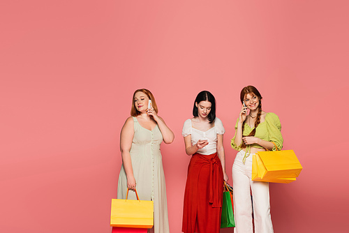 Stylish body positive women with shopping bags talking on smartphones on pink background