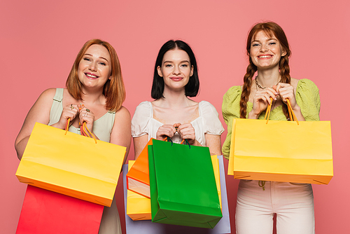 Smiling woman with vitiligo holding shopping bags near body positive friends on pink background