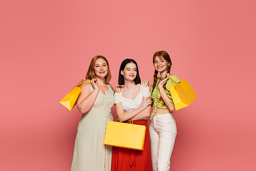 Young body positive women with shopping bags hugging isolated on pink