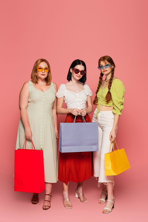 Body positive friends in sunglasses holding shopping bags on pink background