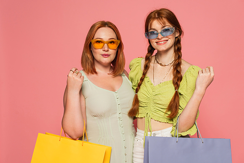 Stylish body positive friends in sunglasses holding shopping bags isolated on pink