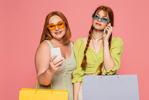 Freckled woman in sunglasses talking on smartphone near friend with shopping bag isolated on pink