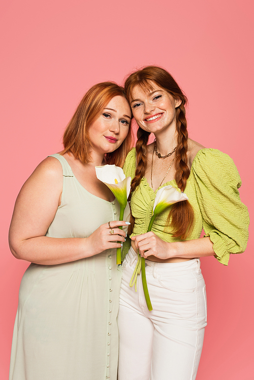 Smiling freckled woman holding calla lily near plus size friend isolated on pink
