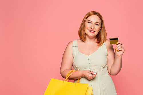 Positive plus size woman holding credit card and shopping bags isolated on pink