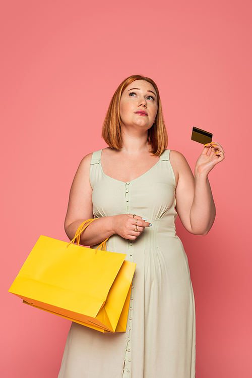 Pensive plus size woman with credit card and shopping bags isolated on pink