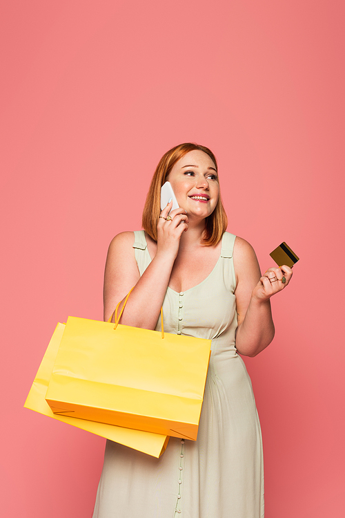 Smiling plus size woman talking on smartphone and holding credit card and shopping bags isolated on pink