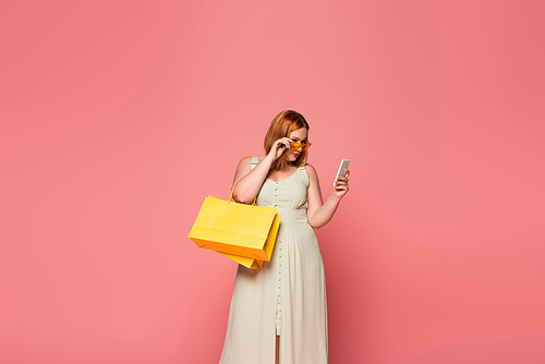 Stylish body positive woman holding shopping bags and smartphone isolated on pink