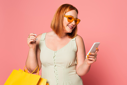 Smiling plus size woman in sunglasses using smartphone and holding shopping bags on pink background