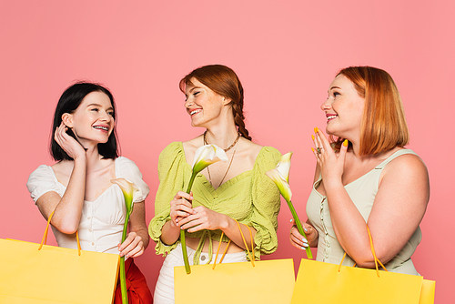 Body positive friends looking at woman with vitiligo and shopping bag with flower isolated on pink