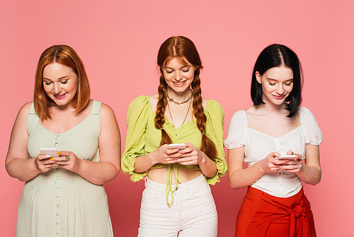 Smiling body positive women using smartphones isolated on pink