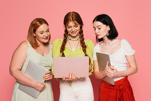 Smiling women looking at friend using laptop on pink background