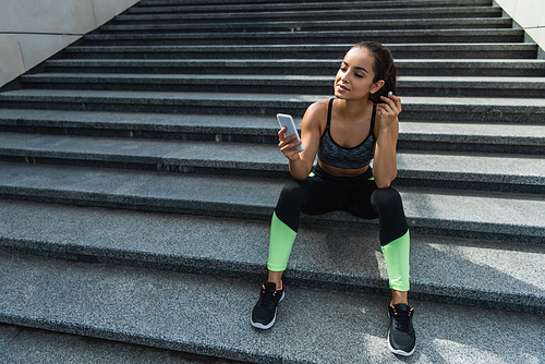 pleased sportswoman in wireless earphones using smartphone while listening music and sitting on stairs outside