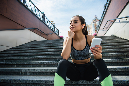 young sportswoman in wireless earphones using smartphone while listening music and sitting on stairs outside