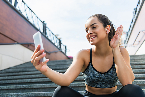 cheerful sportswoman in wireless earphones having video call on smartphone sitting on stairs outside