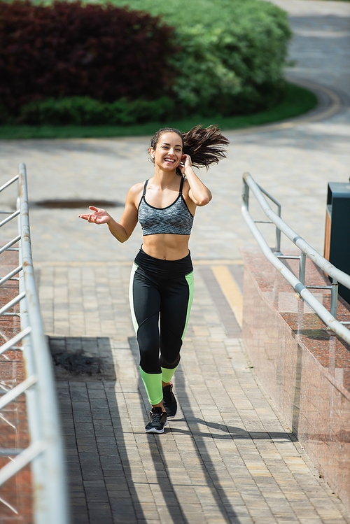 full length of happy woman adjusting wireless earphone while running in park