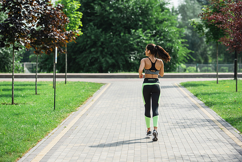 back view of sportive woman in crop top, leggings and wireless earphones listening music while running in park