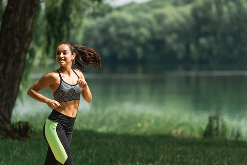 happy sportswoman in wireless earphones listening music while running in green park near lake
