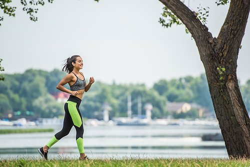 full length of cheerful sportswoman in crop top and leggings jogging in park near lake