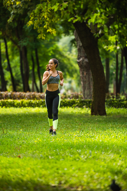 full length of smiling sportswoman in crop top and leggings jogging on grass in park