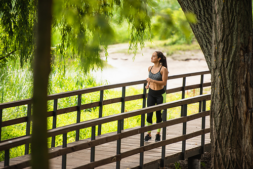 full length of happy sportswoman in crop top and leggings jogging on bridge near lake in park
