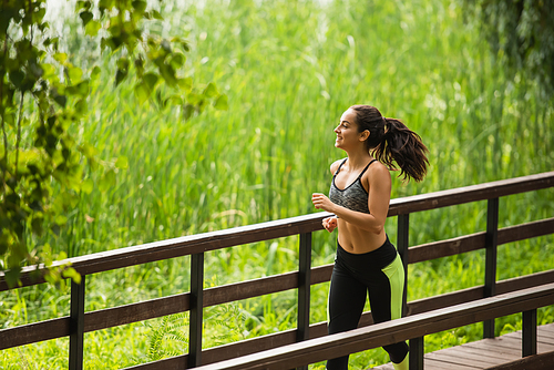 happy woman in crop top and leggings jogging on bridge in green park