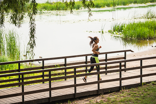 full length of happy woman in crop top and leggings jogging on bridge near lake in park