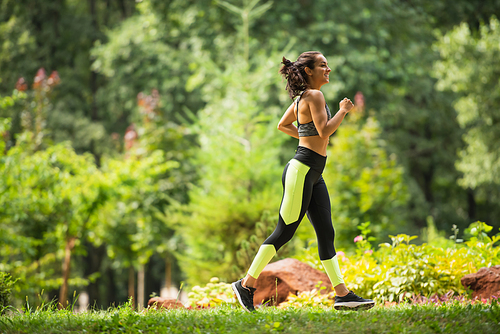 full length of joyful sportswoman in crop top and leggings jogging on grass in park