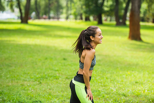 side view of happy athletic woman in crop top resting while standing in green park