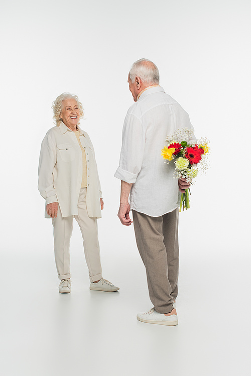 elderly man hiding bouquet of flowers behind back near smiling wife on white