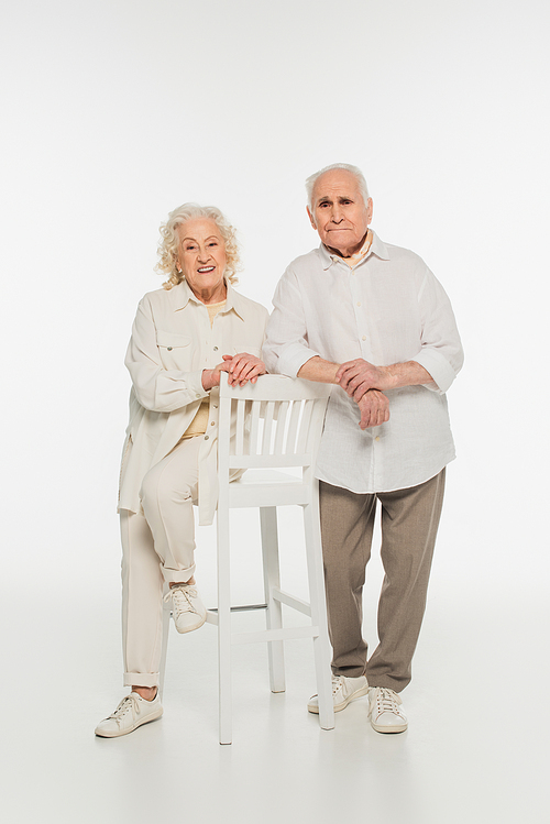 happy elderly couple in casual clothes standing near stool and  on white