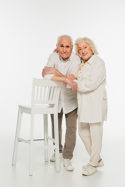 happy elderly couple in casual clothes standing near stool and hugging on white