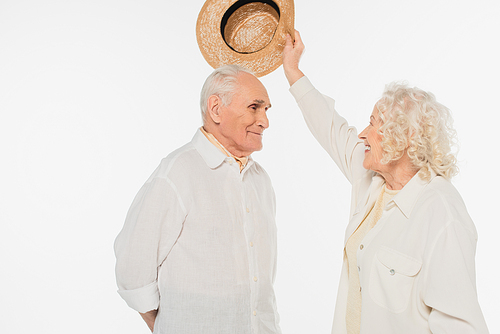 smiling elderly woman putting hat on head of husband isolated on white