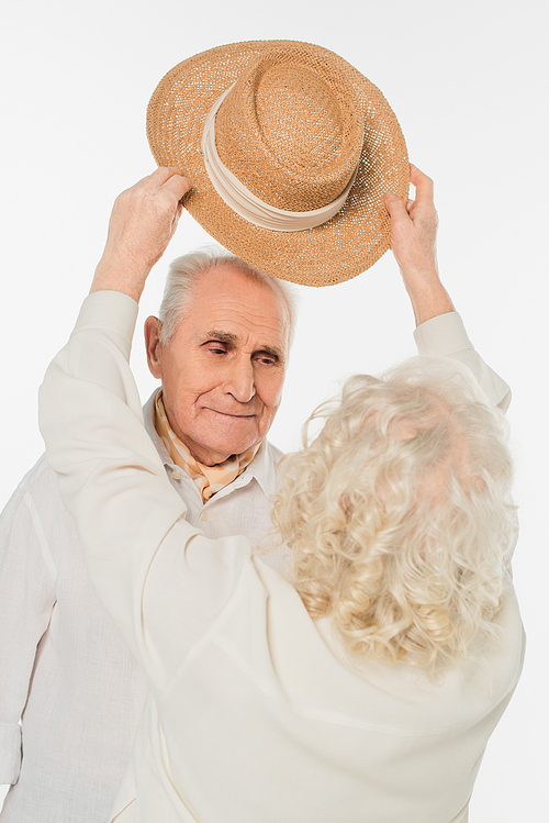 elderly woman putting hat on head of smiling husband isolated on white