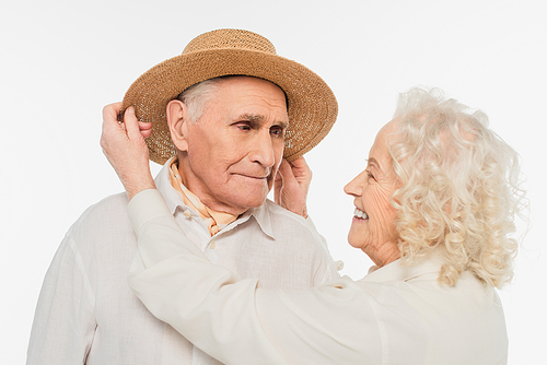 smiling elderly woman adjusting hat on head of husband isolated on white