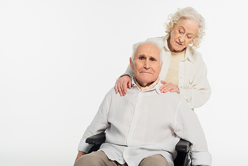 elderly woman holding hands on shoulders of husband in wheelchair isolated on white
