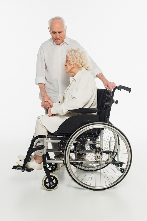 sad elderly woman in wheelchair holding hand of husband on white