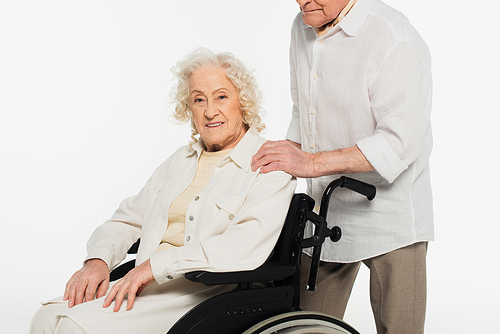 smiling elderly woman in wheelchair near husband holding hand on shoulder isolated on white